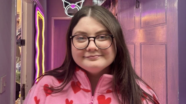 Kobieta z długimi brązowymi włosami i w okularach, ubrana w różową bluzkę z motywem serduszek, stojąca na korytarzu oświetlonym różowym światłem.