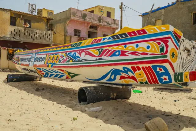 Une pirogue de pêche colorée vue de l'arrière. Elle repose sur des rouleaux sur la plage. On aperçoit quelques bâtiments en bord de mer à l'arrière-plan.