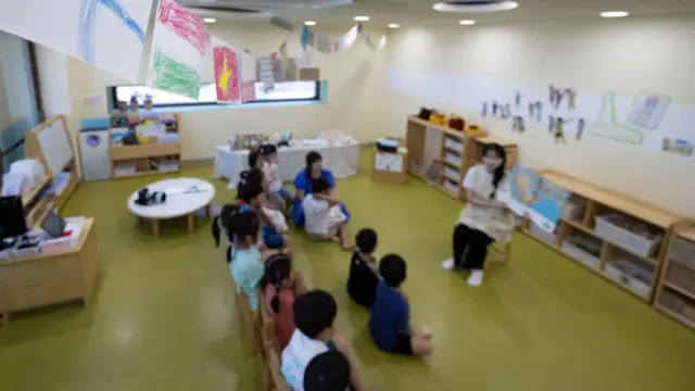 유치원 교실에서 한 교사가 십여명의 아이들 앞에 앉아서 큰 그림 책을 한쪽 팔에 들고 읽어주고 있다 (이 사진은 해당 사건과 관련이 없습니다)