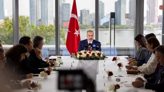 Hakan Fidan'ın arkasında Türk bayrağı var. Uzun bir masada gazetecileri görüyoruz. Arkada fondaysa bir nehir ve gökdelenler var.