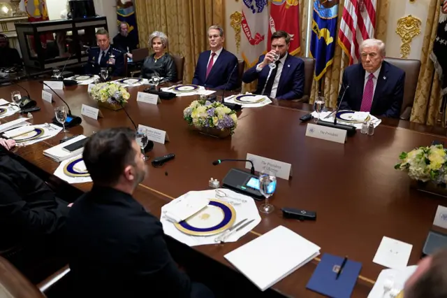 Zelensky, Trump e várias outras autoridades, sentados em torno de uma mesa de madeira na Casa Branca