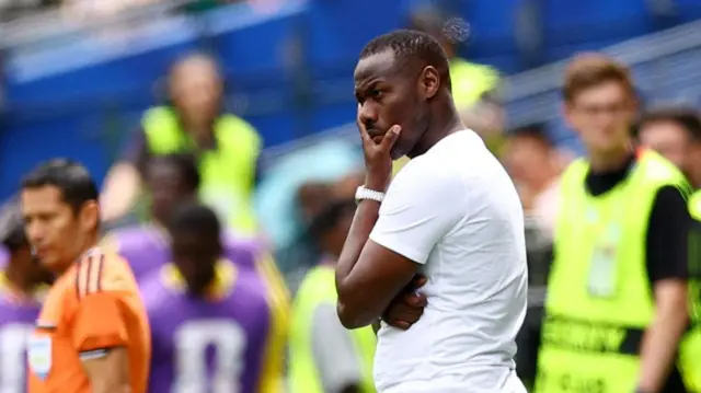 Pape Thiaw, dont on aperçoit la taille jusqu'en haut et qui porte un t-shirt blanc, la main gauche au menton, l'air pensif, sur la ligne de touche pendant un match de football. À l'arrière-plan, flou, on distingue un arbitre assistant en maillot orange, les remplaçants portent des chasubles violettes et les stadiers des chasubles jaune fluo.