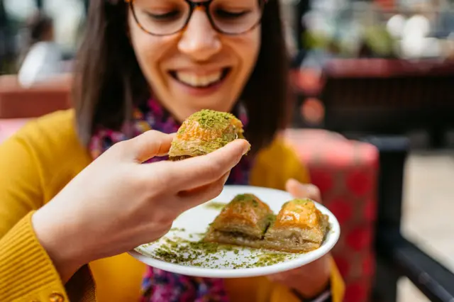 Gözlüklü genç bir kadın gözlerini baklava tabağına dikmiş. Yüzünde heyecan dolu bir ifadeyle tabaktaki üç baklavadan birini ağzına götürüyor. 