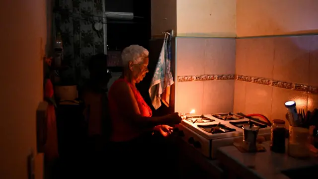 An elderly woman uses a lighter to light her gas stove in order to make coffee. The photograph is dimly lit as electricity has been cut. Picture dated 16 March 2026. 