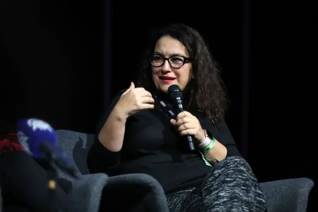 Siyah kazaklı bir kadın. Koltukta oturuyor. Elinde mikrofonla konuşuyor.