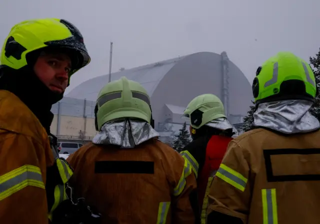 На передньому плані четверо людей у захисному одязі та яскравих касках стоять спиною до камери й дивляться на велетенську аркоподібну споруду. На тлі – велике промислове укриття в туманну, сіру погоду, на даху видно пошкодження та сліди горіння.