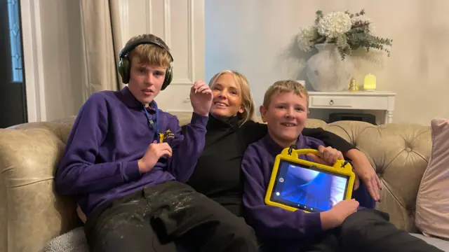 Anne-Marie y sus dos hijos adolescentes autistas sentados en el sofá, sonriendo a la cámara. Anne-Marie lleva un jersey negro, mientras que Tolan y Fredi llevan un uniforme escolar morado.