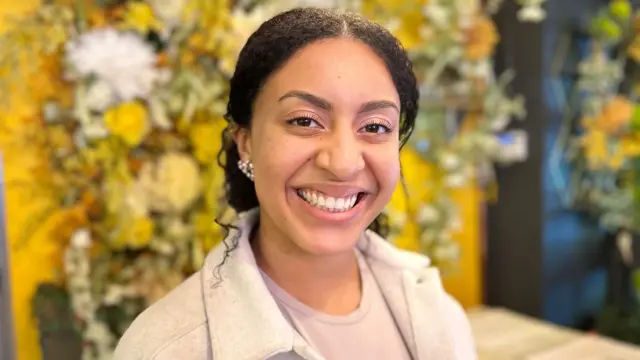 Uma mulher com cabelo preto preso para trás, usando um brinco na orelha direita, uma camisa roxa clara e uma jaqueta branca, está sorrindo em pé em frente a uma parede coberta de flores em um restaurante