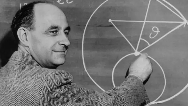 Photo en noir et blanc d'Enrico Fermi, vêtu d'une veste à chevrons, dessinant un diagramme au tableau avec des équations mathématiques.