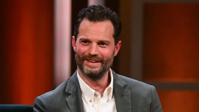 Glumac Džejmi Dornan u beloj košulji i sivom sakou