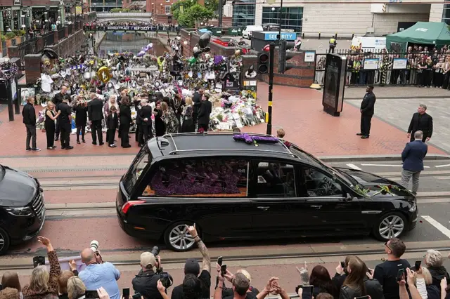 Две черные машины стоят возле фанатского мемориала, вокруг толпы людей.