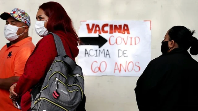 Pessoas de máscara em fila para vacinação. Ao fundo, placa escrito "Vacina Covid Acima de 60 anos"