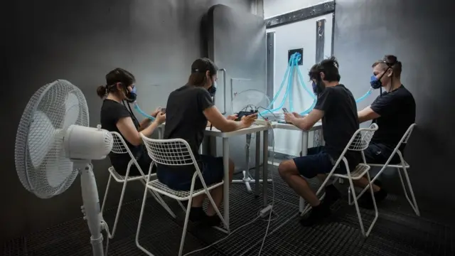 Cuatro personas sentadas alrededor de una mesa vestidas igual, con máscaras de oxígeno en sus bocas.