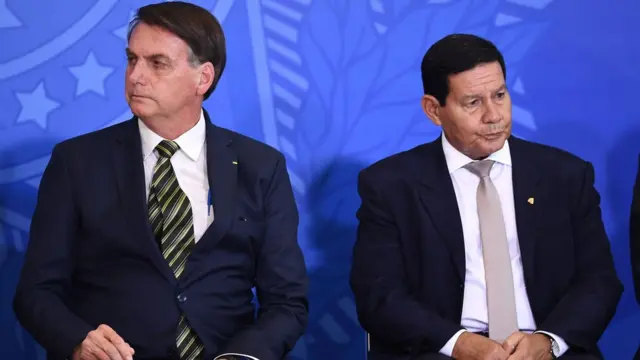 Bolsonaro e Mourão sentados, olhando para lados opostos, em evento
