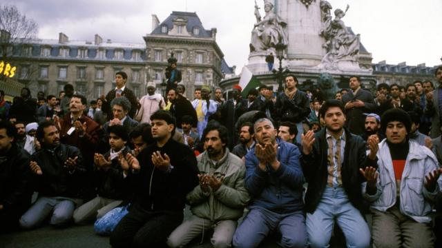 Homens de pele morena clara e cabelos pretos se ajoelham com um prédio e uma estátua parisienses ao fundo