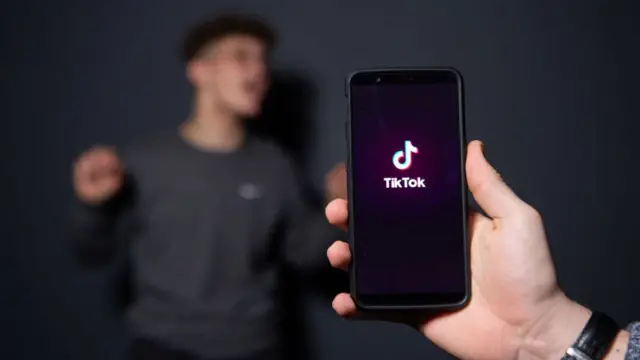 Imagem mostra pessoa segurando smartphone com a logo do TikTok aparecendo na tela e outra pessoa, ao fundo, sendo filmada