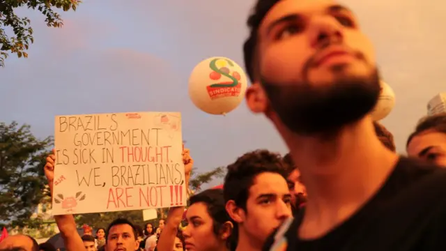 RostosmanifestantesSão Paulo, entre os quais um que segura placa dizendoinglês: 'O governo brasileiro é doente no pensamento; nós, brasileiros, não'