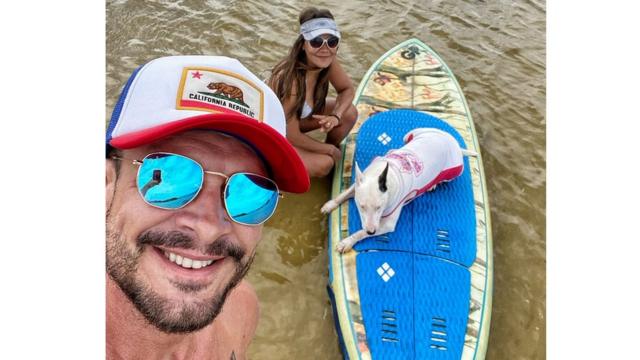 Andreza e o marido sorriem para selfie, ao lado da cachorra de estimação em cima de prancha na água