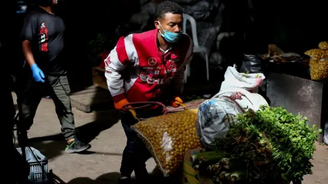 Un hombre con una mascarilla lleva una carretilla con verduras.