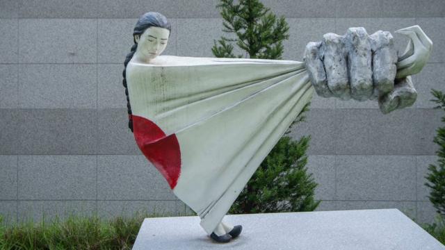 Estatua de una mujer siendo apresada por una bandera japonesa.