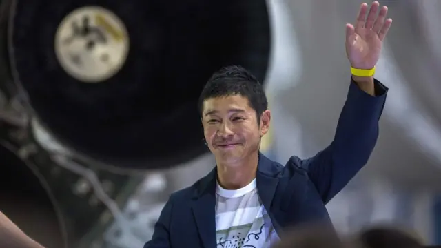 Yusaku Maezawa sorrindo e com a mão esquerda erguida, cumprimentando o público