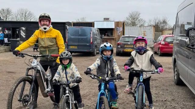 Ryan Horsfall y sus tres hijos pequeños, todos en bicicleta y con casco, sonríen a la cámara.