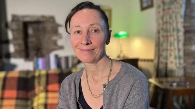 Dr Hard sitting on her sofa wearing a grey cardigan and gold necklace