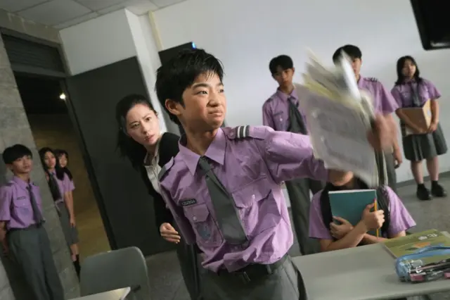 《自杀通告》剧照，一名身穿紫色校服的男学生神情激动，身后有一名女子拉着他的手臂，后方有多名同样穿着紫色校服的学生观看着。