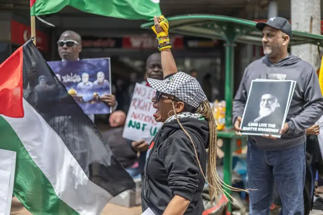Une femme vêtue d'un sweat à capuche noir, d'une casquette noire et blanche et de gants jaunes lève le poing en l'air. Derrière elle, des manifestants brandissent des pancartes sur lesquelles figure une photo de l'ayatollah Khamenei, guide suprême de l'Iran.