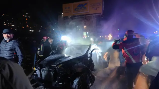 Patlamış bir araç ve çevresinde kalabalık görünüyor, gece saatleri. 