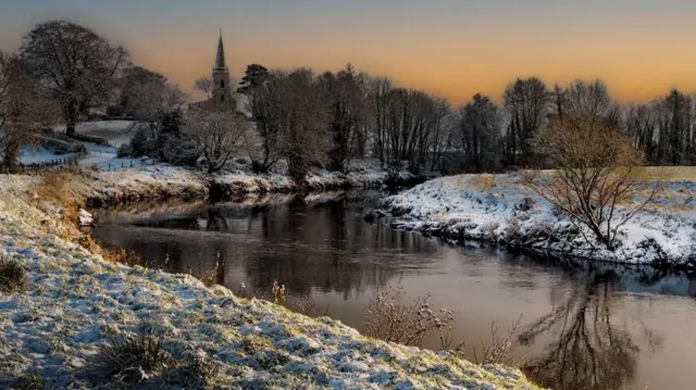 Un río con nieve en las orillas y una iglesia y árboles a lo lejos. El cielo es naranja.