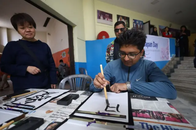 नेपाल र जापानबीच शिक्षा र कलासहितका क्षेत्रमा आदानप्रदान हुँदै आएको छ। तस्बीरमा निलो रङ्गको ज्याकेट लगाएका एक युवाले जापानी अक्षर लेखिरहेको देख्नसकिन्छ। उनको छेउमा निलो स्वेटर लगाएकी एक महिला र पृष्ठभूमिमा एक जना पुरुष उभिइरहेका छन्।