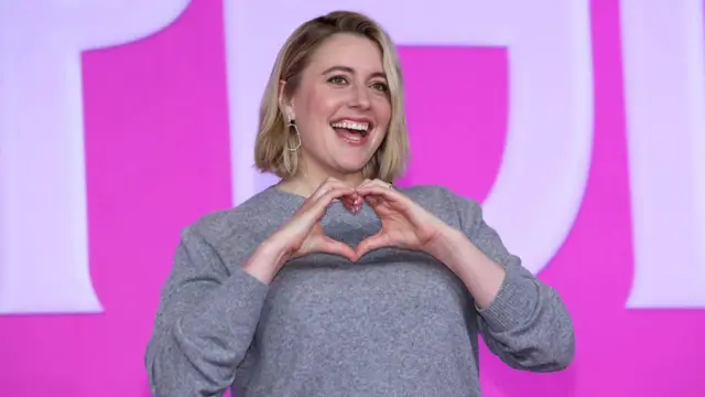 Greta Gerwig sorrindo e fazendo coração com as mãos