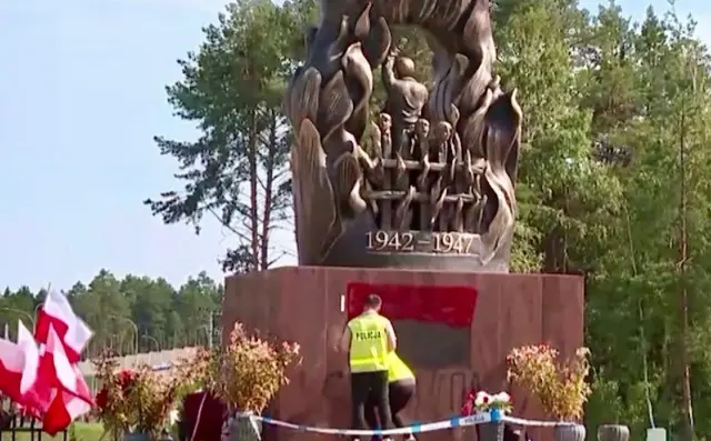 Пам’ятник із зображенням групи людей у вогняному полум’ї, з датами "1942–1947" на постаменті. Перед монументом двоє поліцейських працюють за обмежувальною стрічкою. На плиті памʼятника фарбою намальований червоно-чорний прапор. Ліворуч видно кілька польських прапорів і квіти.