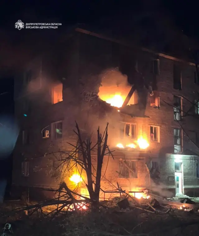 Багатоповерховий житловий будинок, охоплений вогнем. У верхній частині фасаду утворився великий пролом, у кількох квартирах вибиті вікна та пожежа. Біля будинку помітні уламки й пошкоджені дерева, територію огортає дим.