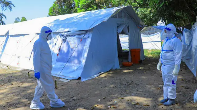 ሰውነትን በሙሉ የሚሸፍን ነጭ የደህንነት መጠበቂያ የለበሱ ሁለት የጤና ባለሙያዎች ከነጭ ድንኳን ፊት ለፊት ይታያሉ