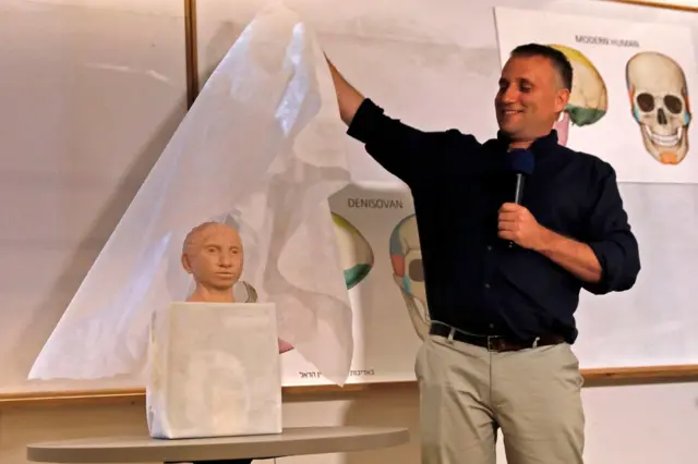 El profesor Liran Carmel, con una camisa negra y un pantalón beige, levanta una cubierta blanca y deja al descubierto el modelo de un rostro beige