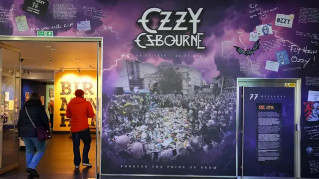 Dos personas pasan junto a un puesto de museo que exhibe la exposición de Ozzy Osbourne, que muestra su desfile fúnebre y sus homenajes. Una visitante lleva un abrigo negro y vaqueros, y el hombre, un abrigo rojo, un sombrero rojo y vaqueros.