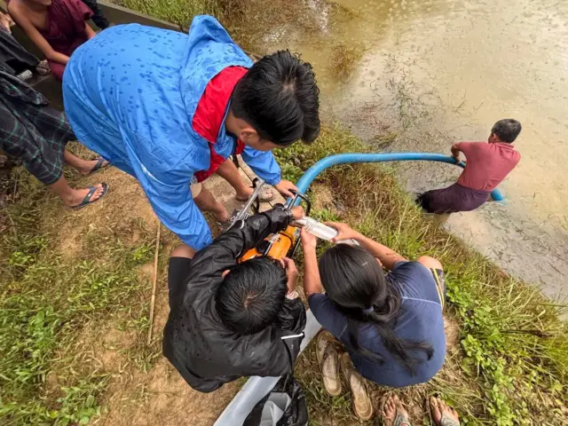 ရေကြီးလို့ ဝါကျင့်ကျင့်ဖြစ်သွားတဲ့ ရေကန်ထဲကို ရေပိုက်ကြီးနဲ့ မီးစက်ကိုသုံးကာ ပြန်လည် သန့်စင်နေကြတဲ့ အမျိုးသားလေးဦး။ အင်္ကျီအနီရောင်ဝတ် အမျိုးသားက ကန်ထဲက ပိုက်ကို ကိုင်ထားပြီး လူငယ်အမျိုးသား ၃ ဦးကို မီးစက်နှိုးဖို့ ရေထည့်နေတဲ့မြင်ကွင်း