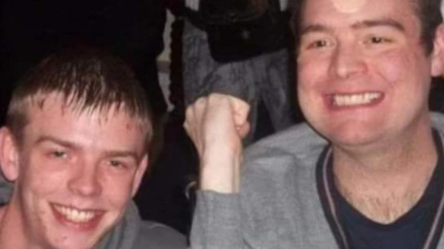 Dos hombres vestidos con suéteres grises, ambos sonrientes, uno con cabello rubio oscuro y otro con cabello castaño oscuro, levantando la mano en señal de celebración.