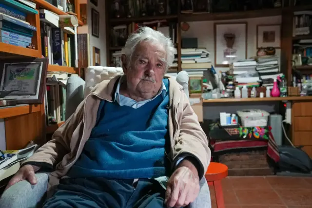 José Mujica, um homem idosoroleta com multiplicadorcabelos brancos e bigode, vestido com camisa azul e casaco bege, olhando para a câmera. Ao fundo, um escritório repletoroleta com multiplicadorlivros e outros objetos