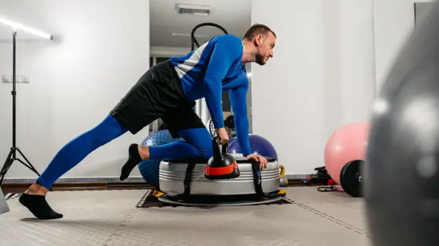 Młody, umięśniony mężczyzna trenuje na siłowni na platformie power plate z odważnikiem kettlebell.
