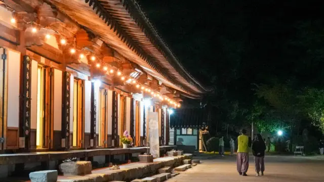 Bangunan kuil tradisional Korea yang diterangi cahaya hangat pada malam hari, dengan atap genteng melengkung, tiang-tiang kayu, dan tangga batu yang membatasi halaman luas. Beberapa pengunjung berjalan di sepanjang jalur di samping kuil tersebut.