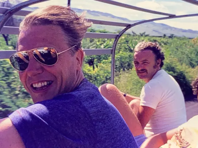 Attenborough y otro hombre sentados en un vehículo abierto recorriendo un paisaje verde y montañoso; el famoso documentalista está en primer plano con gafas de sol