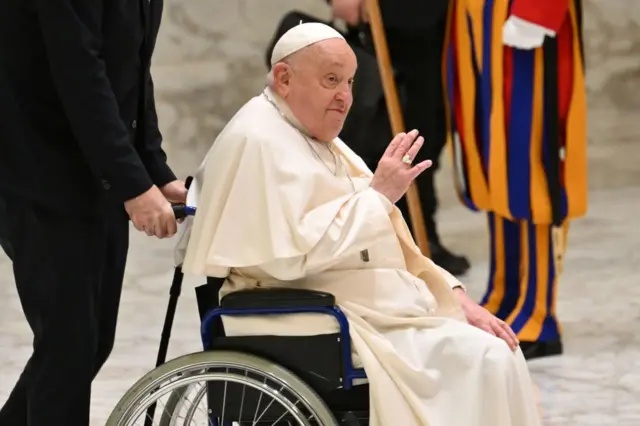 Papa Francisco acenando enquanto é conduzido em cadeira de rodas