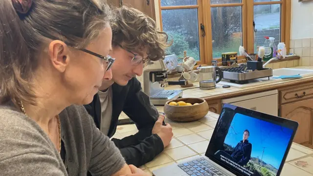 Dr Bernadette Hard and son Callum in her kitchen watching a documentary filmed Callum on a laptop