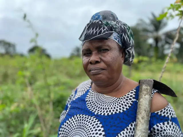 Ledisi Nomkia get a hoe over her shoulder as she look into di camera. For di background na farm.