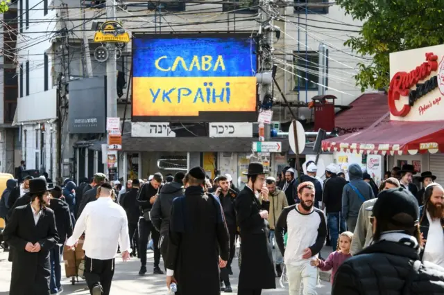 Десятки єврейських паломників у традиційному вбранні на вулиці Умані. На великому екрані – український прапор із написом "Слава Україні", поруч кілька вивісок їдишем