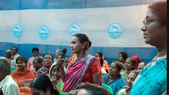 Mulher de pé fala ao microfone em meio a uma reunião comunitária, rodeada de diversas pessoas sentadas em cadeiras de plástico, com um fundo azul e branco com logotipos circulares