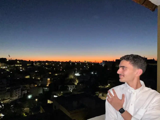 صورة لمحمد أديب (21 سنة) طالب إدارة، يرتدي قميص أبيض وتظهر خلفه مدينة عمّان